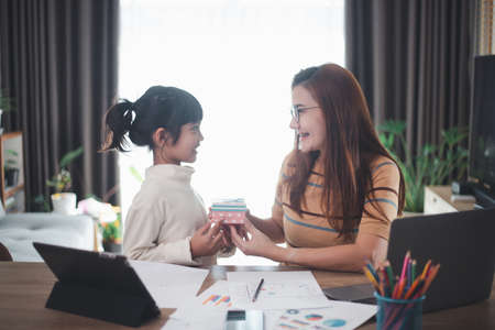 Happy Mother's day. Young woman mom getting congratulations from excited cute little daughter at home, child giving mother gift box while she working on laptop. Family holidays conceptの写真素材