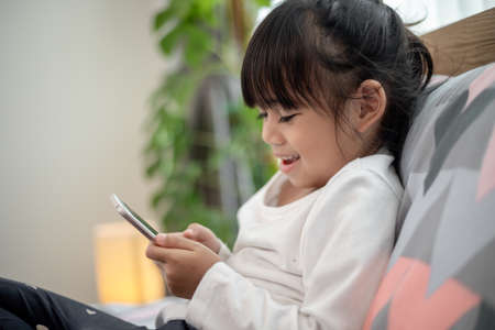 Cute little kid girl watching video on smartphone with smiley face alone on the bed, child using mobile phone with happy face at home. Stay at home quarantine coronavirus COVID-19 pandemic prevention.の写真素材