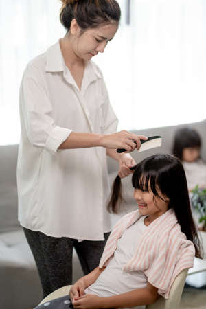 Asian Mother cutting hair to her daughter in living room at home while stay at home safe from Covid-19 Coronavirus during lockdown. Self-quarantine and social distancing concept.の写真素材