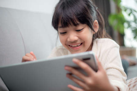 Little girl using tablet playing game on the internet, Kid sitting on sofa watching or talking with a friend online, Child relaxing in living room in the morning, Children with New Technology conceptの写真素材