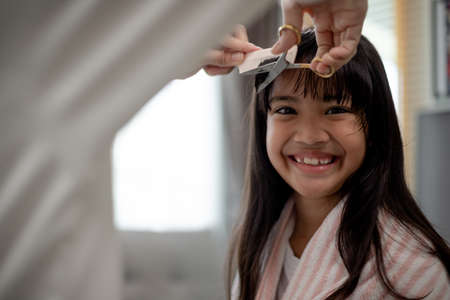 Asian Mother cutting hair to her daughter in living room at home while stay at home safe from...