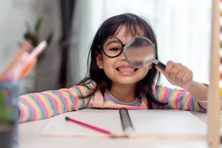 Student little Asian girl writing with a magnifying glass looking at the camera. sweet kids sitting...