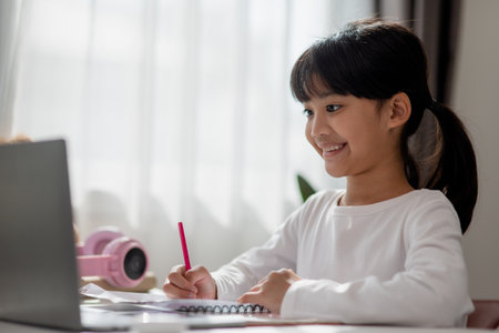 Asian schoolgirl doing her homework with laptop at home. Children use gadgets to study. Education and distance learning for kids. Homeschooling during quarantine. Stay at homeの写真素材