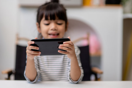 Cute little girl uses smartphone while sitting at the sofa in the living room. Child surfing the internet on mobile phone, browses through internet and watches cartoons online at homeの写真素材