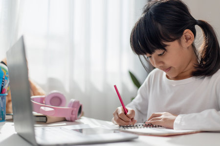 Asian schoolgirl doing her homework with laptop at home. Children use gadgets to study. Education and distance learning for kids. Homeschooling during quarantine. Stay at homeの写真素材