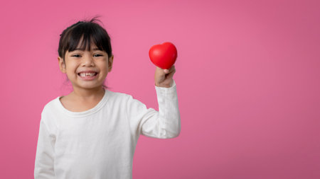 Little Asian girl holding red heart shaped application and looking at camera happily on pink.の写真素材