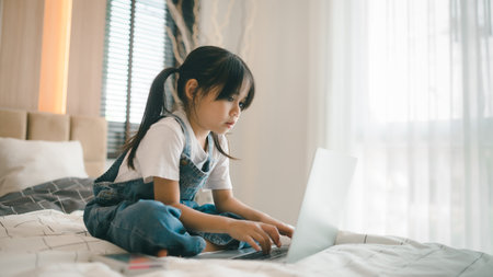 Asian girl child sitting in bed and learning online on laptop Internet. Virtual class lesson on video during self isolation at home. Distant remote video education. Modern school study for kidsの写真素材