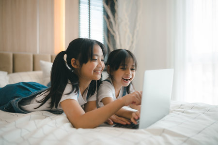 Sibling Asian girl child on the bed and learning online on a laptop Internet. Virtual class lesson on video during self isolation at home. Distant remote video education. Modern school study for kidsの写真素材