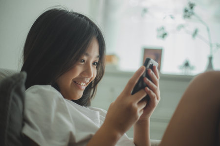 Happy Asian girl relaxing on a comfortable couch at home texting and messaging on a smartphone, smiling girl uses a cellphone, chatting online message, and shopping online from home.の写真素材