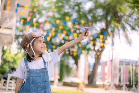 A young girl wearing a pilot's hat and a white shirt is holding a paper airplane. She is smiling and...