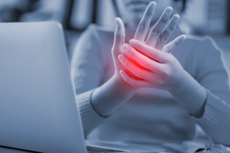 A woman is holding her hand up in pain while sitting in front of a laptop. The laptop is open and...