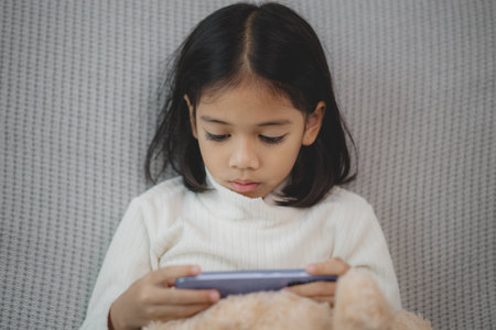 A young girl is playing a video game on a tablet while sitting on a bed. She is wearing a white...