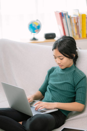 Asian young girls doing homework on laptops on a comfortable couch.