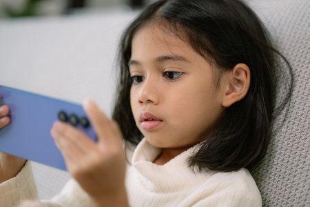 A young girl is sitting on a couch and playing a video game on a cell phone. She is focused on the...