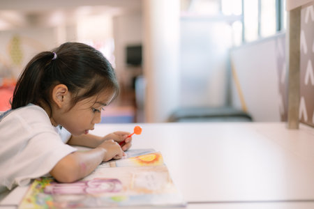 A young girl is sitting at a table reading a book. She is holding a red and orange toy in her hand
