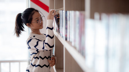 A young girl is looking through a library shelf. She is wearing glasses and a striped shirt. The scene is set in a library with many books on the shelvesの写真素材