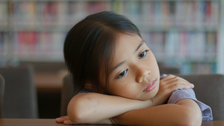 An Asian girl sits at a table in a library. She looks bored and lost in thought. Her disinterested expression shows her disinterest in the things around her.の写真素材