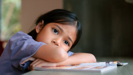 An Asian girl sits at a table in a library, her face clearly showing boredom as she looks away from the open book in front of her. She looks disinterested and lost in thought.の写真素材