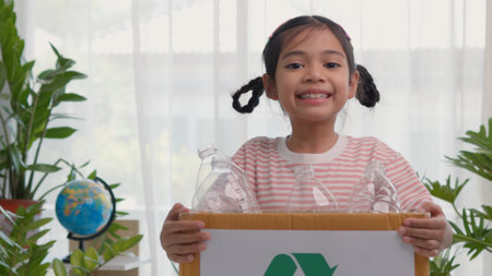 A cute Asian girl carries a paper box full of plastic water bottles. Children sort out trash for recycling. Cultivate the learning of waste separation. The child smiles sweet and brightの写真素材