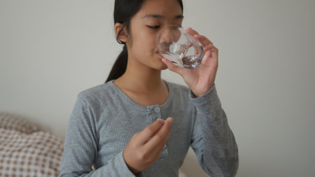 Have a fever, Asian girl hand taking medicine, a pill for a headache, stomach pain or take vitamins holding a glass of water sitting in a bedroom at home. Healthcare concepts.の写真素材