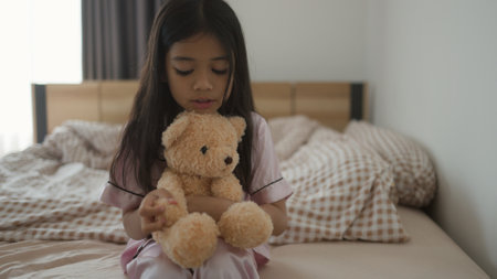 Sweet Asian little girl is hugging a teddy bear and smiling while sitting on her bed at homeの写真素材