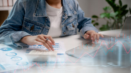 Close-up of a woman using a calculator and laptop with financial graphs overlay, concept of data analytics, stock trading, digital investment, and business financial planning.の写真素材