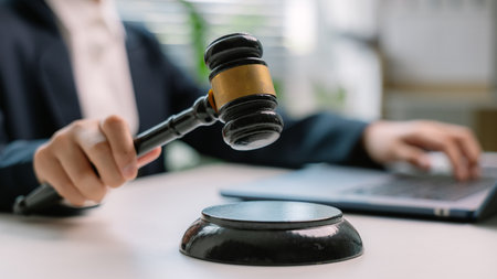 Close-up of judge's hand holding gavel with laptop in background, representing legal decision,...