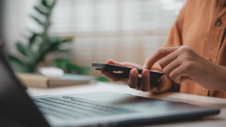 Close-up of person using smartphone while working on laptop, representing digital communication, mobile productivity, smart office, multitasking, and remote work lifestyle.の写真素材