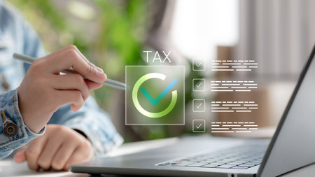 Hand holding pen checking online tax form on laptop with digital checklist. Concept of e-filing, tax return, and remote tax preparation for individuals or businesses.の写真素材
