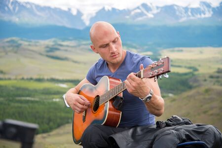 bald motorcyclist plays guitar sitting on a motorcycle on the background of mountainsの写真素材