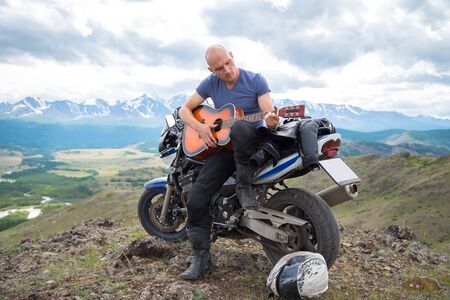 bald motorcyclist plays guitar sitting on a motorcycle on the background of mountainsの写真素材