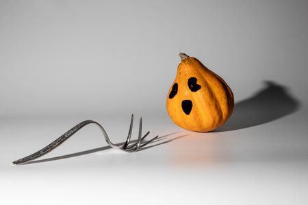 Conceptual Halloween photograph made from an orange ornamental pumpkin and a deformed stainless steel fork, with prominent shadowsの写真素材