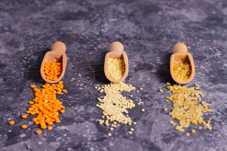 Raw orange lentils, millet and bulgur for proper nutrition and health in wooden spoons on a gray...