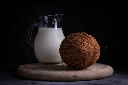 Whole brown coconut with a jug of coconut vegan milk on a wooden stand on a gray background in low keyの写真素材