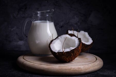 Chopped ripe brown coconut with a jug of coconut vegan milk on a wooden stand on a gray background in low keyの写真素材