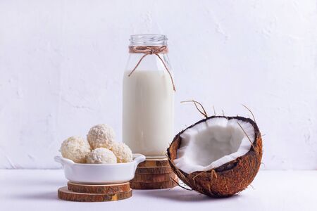 Bottle of coconut vegan milk on wooden coasters with chopped brown coconut and round sweets with...