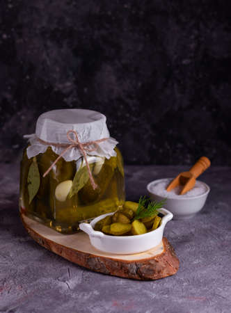 Fermented canned cucumbers with bay leaf, garlic and dill in a glass jar with a white plate with sliced cucumber on a gray backgroundの写真素材