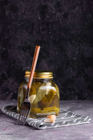 Fermented canned cucumbers with bay leaf, garlic and dill in a glass jar with a fork on a gray backgroundの写真素材