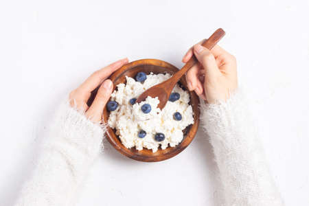 Girl in a white sweater holds a plate with grained cottage cheese and blueberries in a spoon