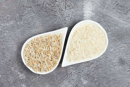 Brown uncooked and white round rice in white ceramic plates on gray backgroundの写真素材