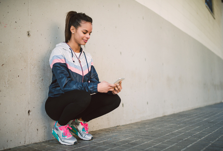 Hispanic runner woman squatting and looking her smart phoneの写真素材
