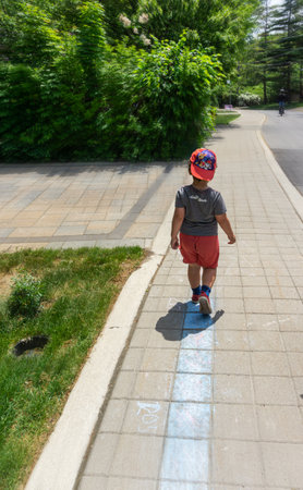Toronto, Canada, July 2021 - Little boy wearing shorts and cap walking alone on the sidewalk of a residential neighbourhood on a bright summer dayのeditorial素材