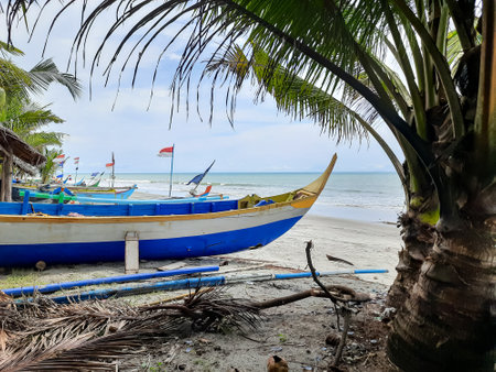 Traditional Indonesian fishing boats docked on a sandy beach, surrounded by palm trees and...