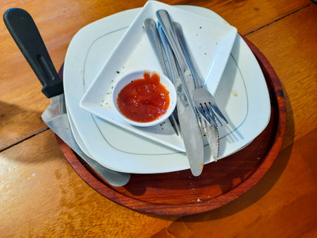 A stack of used plates with cutlery and a small bowl of leftover tomato sauce on a wooden table, showing the aftermath of a finished mealの写真素材