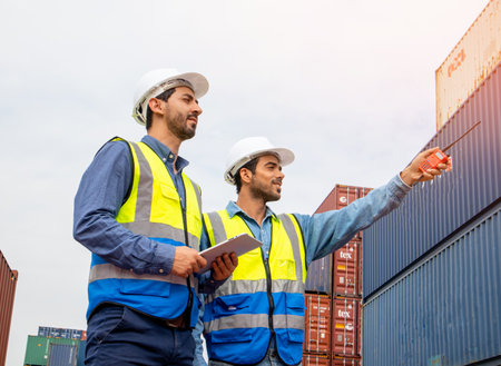 Two professional engineers in safety helmets and uniforms consulting each together in a container...
