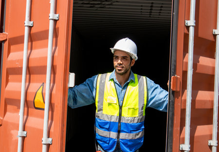 A male worker wearing a safety vest and helmet opens the container door and conducts regular...