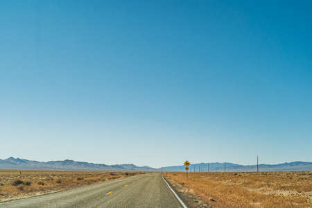 Two lane desert highway road leading to the horizon against clear blue sky and mountainsの写真素材