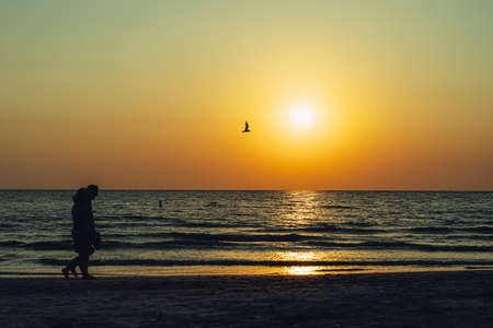 Two people walk along the beach in silhouette at sunset against orange skyの写真素材
