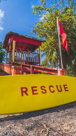 Red Lifeguard tower and yellow rescue board with red warning flag waving in light breeze against blue sky. low angle perspectiveの写真素材