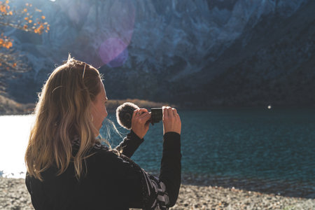 Female travel influencer filming with video camera in front of mountain lake